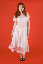 A woman with red hair wearing a white lace and mesh midi dress with short sleeves and black high heels, standing against a solid red studio background.