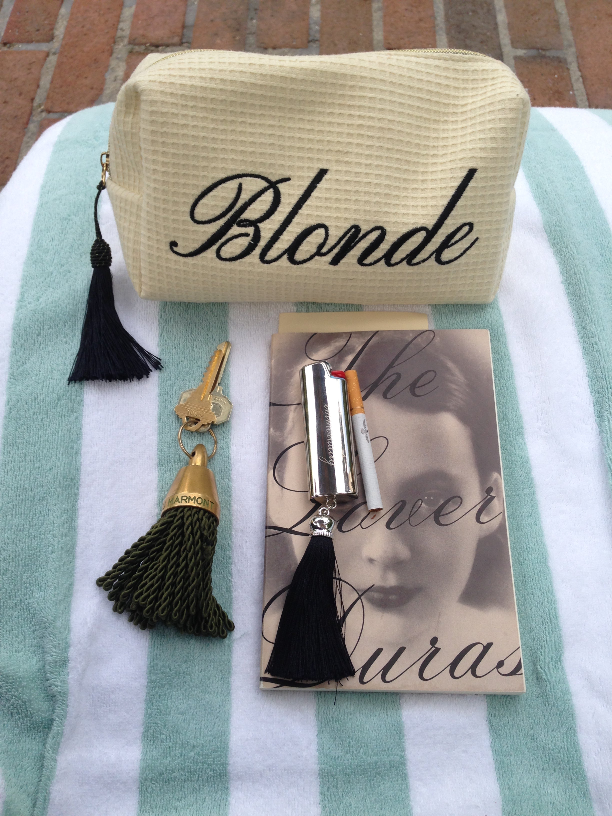 A beige waffle-weave cosmetic pouch with 'Blonde' written in black script, displayed on a mint and white striped towel alongside a black tassel keychain, a gold key, a silver lip product, and a book.