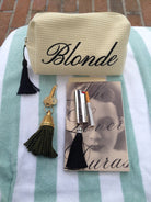 A beige waffle-weave cosmetic pouch with 'Blonde' written in black script, displayed on a mint and white striped towel alongside a black tassel keychain, a gold key, a silver lip product, and a book.