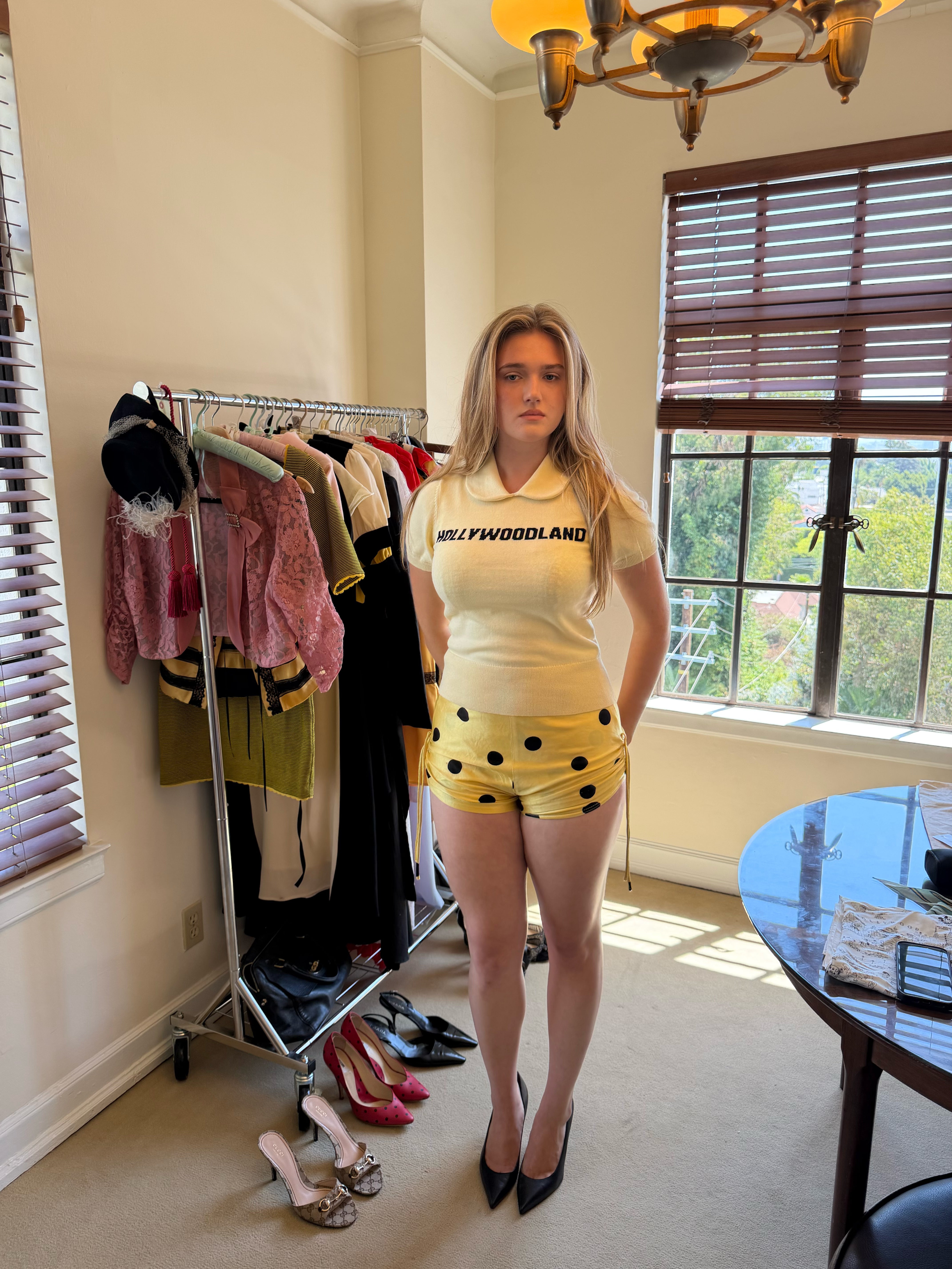 A woman stands in a room wearing a pale yellow short-sleeve polo shirt with 'HOLLYWOODLAND' printed across the chest and matching yellow shorts with black polka dots. She is posed in front of a clothing rack filled with other garments and shoes, with windows and blinds behind her.