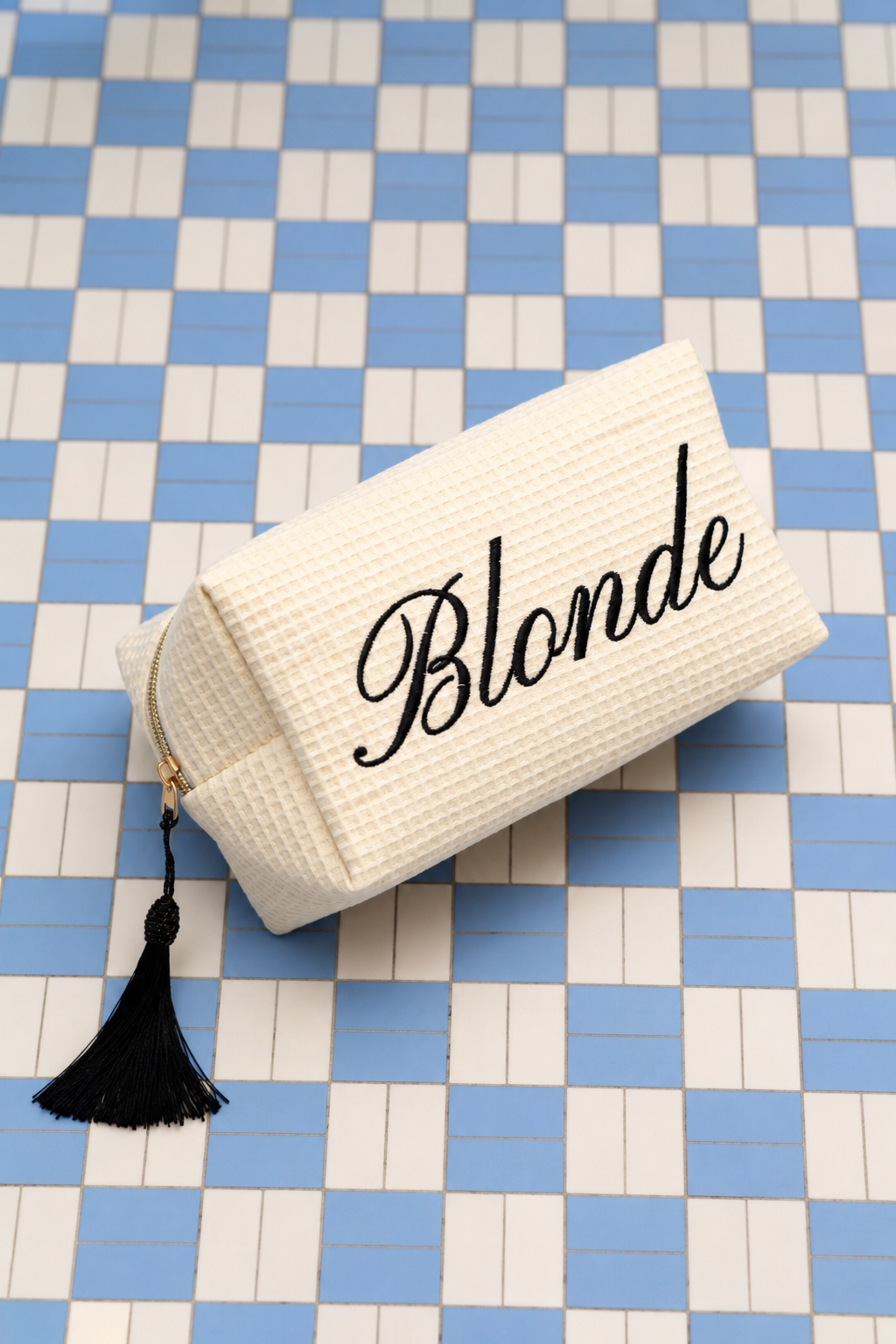 A rectangular cream-colored waffle-weave makeup pouch with a black script 'Blonde' print on the front, resting on a blue and white checkered tile surface. The pouch has a black tassel zipper pull.